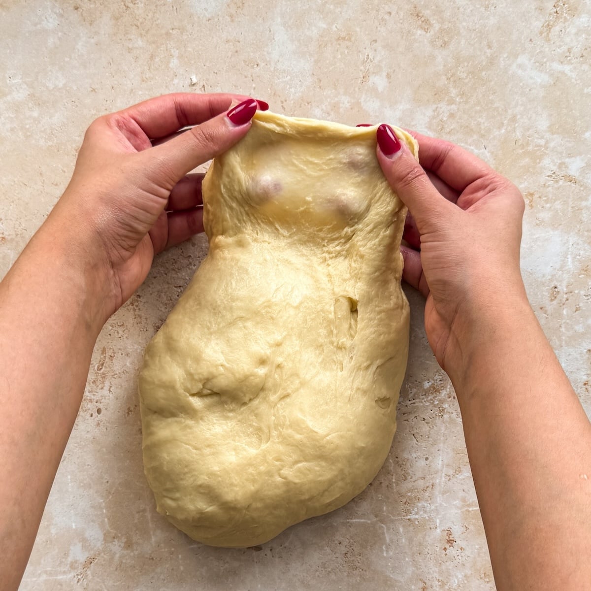 Two hands with red nail polish stretch the top edge of a ball of dough on a light countertop, revealing its elasticity