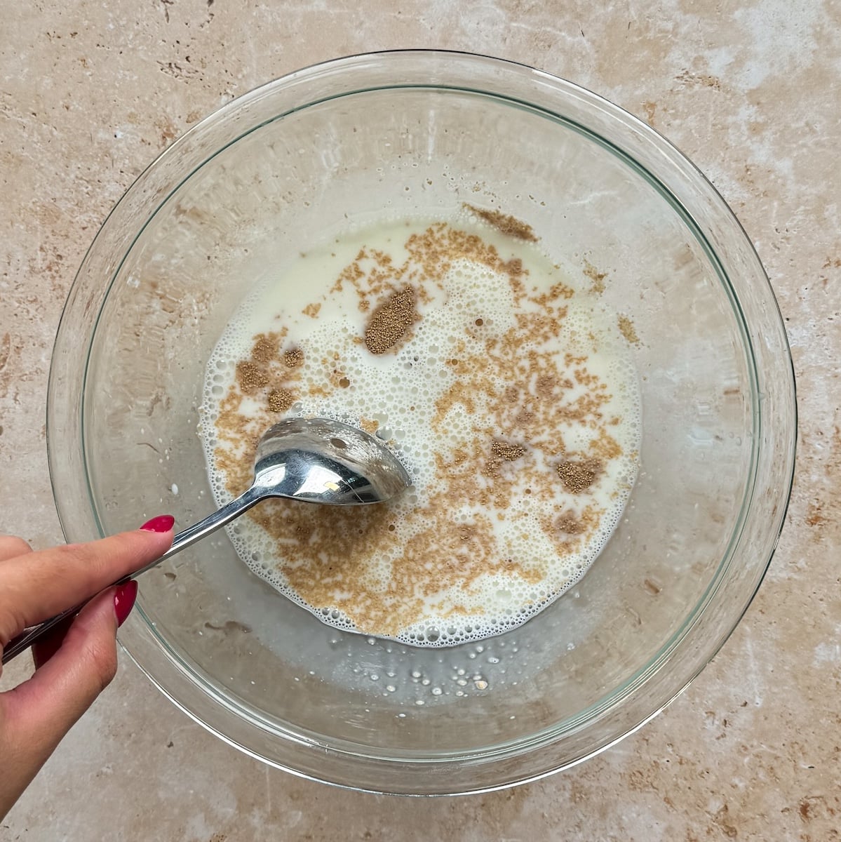 A hand holding a spoon stirs a mixture of yeast, milk, and sugar in a large clear glass bowl on a beige countertop
