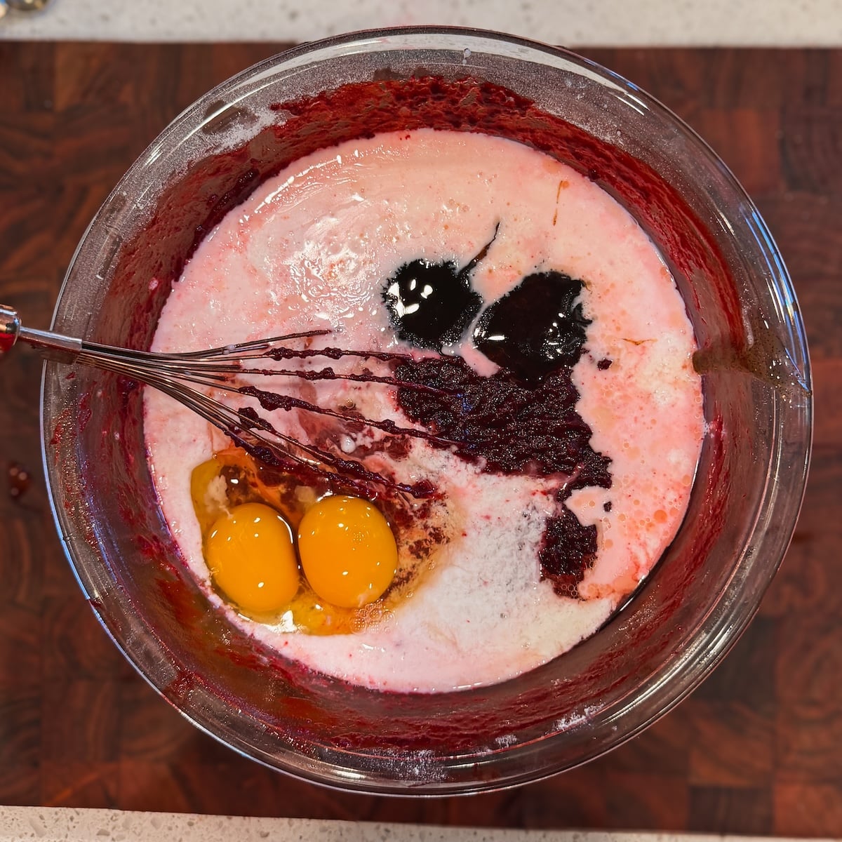 A glass bowl with two cracked eggs, cake batter, milk, and other baking ingredients being whisked together, viewed from above on a wood cutting board