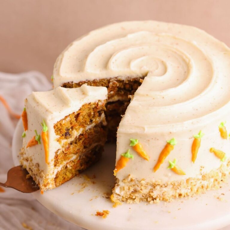 carrot cake on a cake stand with a spatula pulling out a slice. the cake is covered in brown butter cream cheese frosting and decorated with mini frosting carrots and walnuts. The cake looks very moist and loaded