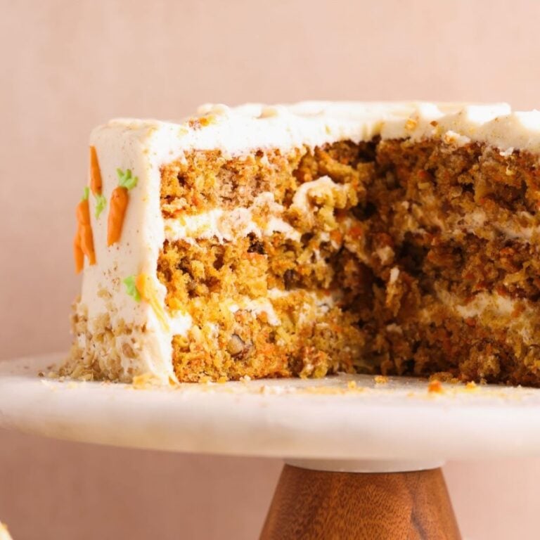 carrot cake on a cake stand with a spatula pulling out a slice. the cake is covered in brown butter cream cheese frosting and decorated with mini frosting carrots and walnuts. The cake looks very moist and loaded. close up picture from the side