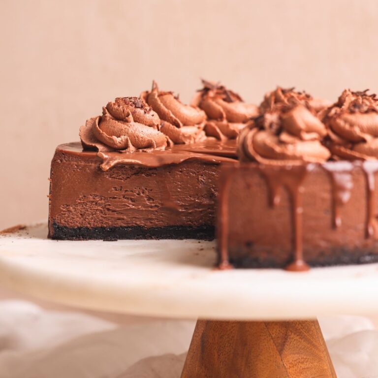 chocolate cheesecake on a marble board with a few slices removed to show the cross-section. the cheesecake has a crispy oreo crust, a rich and creamy texture, and is decorated with ganache and chocolate whipped cream. picture from the side