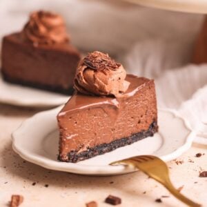 slice of chocolate cheesecake on a small plate with another in the background. the cheesecake has a crispy oreo crust, a rich and creamy texture, and is covered with ganache and a dollop of chocolate whipped cream