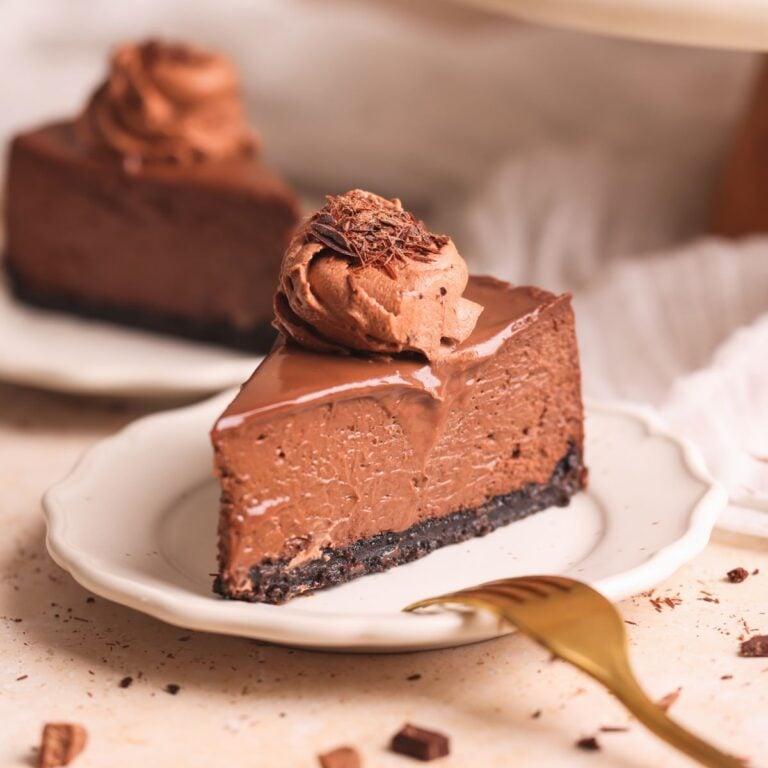 slice of chocolate cheesecake on a small plate with another in the background. the cheesecake has a crispy oreo crust, a rich and creamy texture, and is covered with ganache and a dollop of chocolate whipped cream