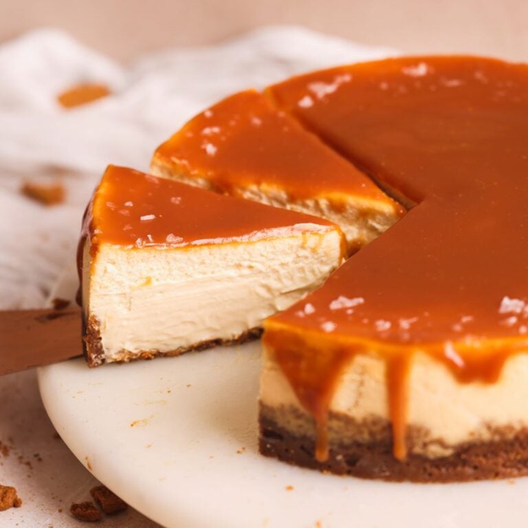 salted caramel cheesecake on a marble board. cheesecake has been drizzled with salted caramel sauce and decorated with sea salt flakes. A slice has been removed and another is being taken out by a spatula, showing the extra creamy interior