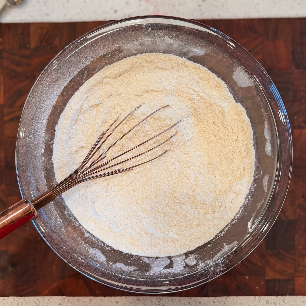 A glass bowl filled with flour and a metal whisk with a red handle sits on a wooden surface, ready for baking preparation
