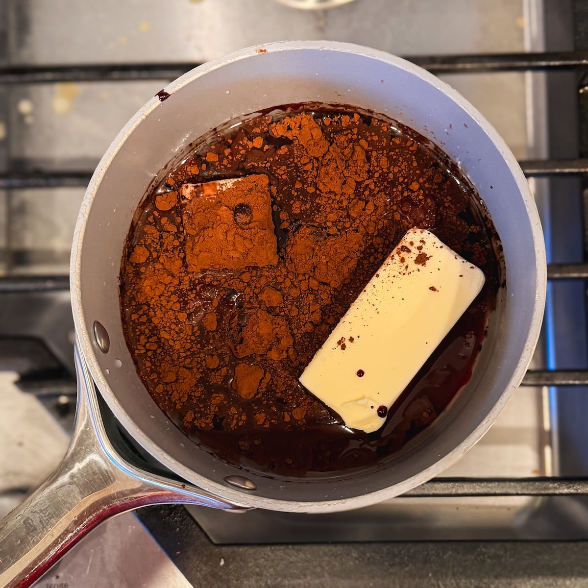 A saucepan on a stove contains butter, cocoa powder, oil and water, being heated together