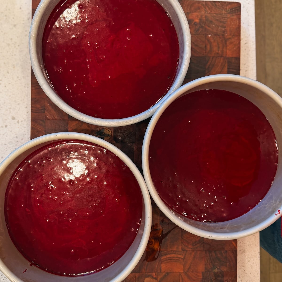 Three round cake pans filled with smooth, bright red batter are placed on a wooden cutting board
