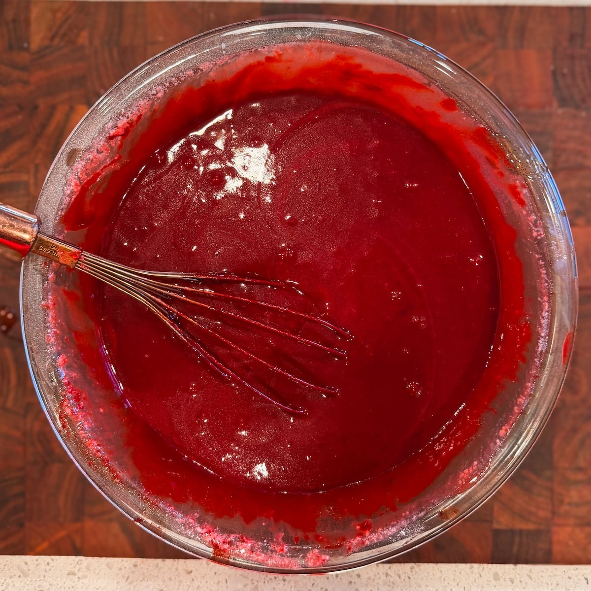 A glass bowl filled with glossy red cake batter is being mixed with a metal whisk. The bowl rests on a dark wooden surface.