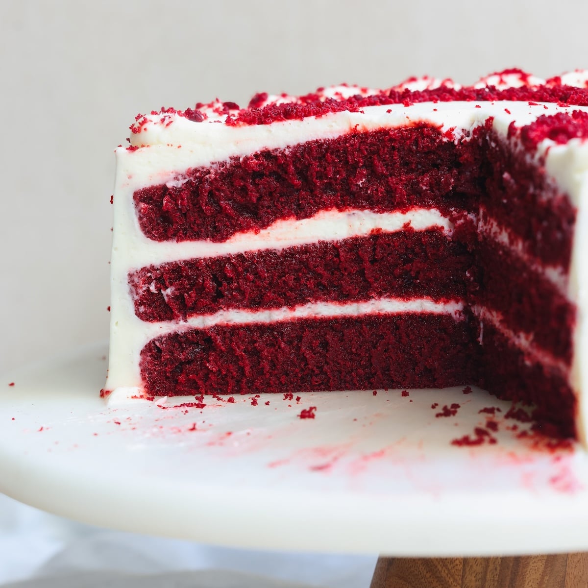 A three-layer red velvet cake with white cream cheese frosting sits on a white cake stand, with a large slice missing, revealing the cake's rich and moist red layers and smooth icing.
