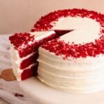 red velvet cake on a marble cake stand with a spatula pulling out one slice. The cake is covered in cream cheese frosting and decorated with red velvet cake crumbs. Picture from an angle