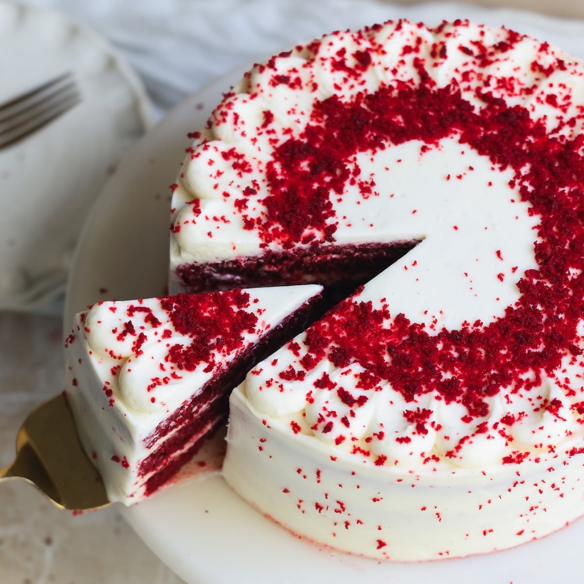 A red velvet cake with cream cheese frosting and red crumbs on top. A single slice is being lifted from the cake on a white cake stand.