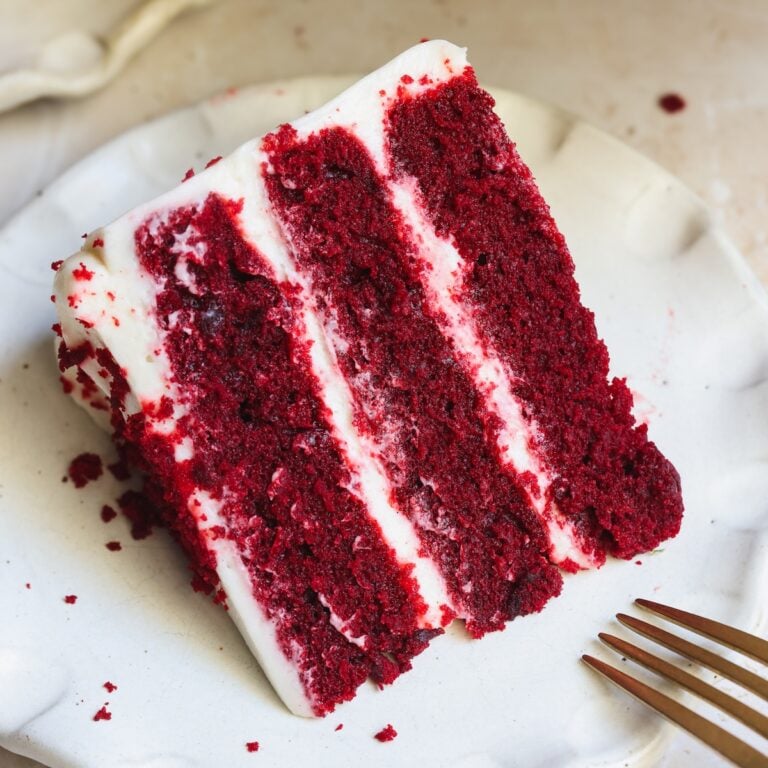 A slice of red velvet cake, with three layers and cream cheese frosting, sits on a white plate next to a fork.