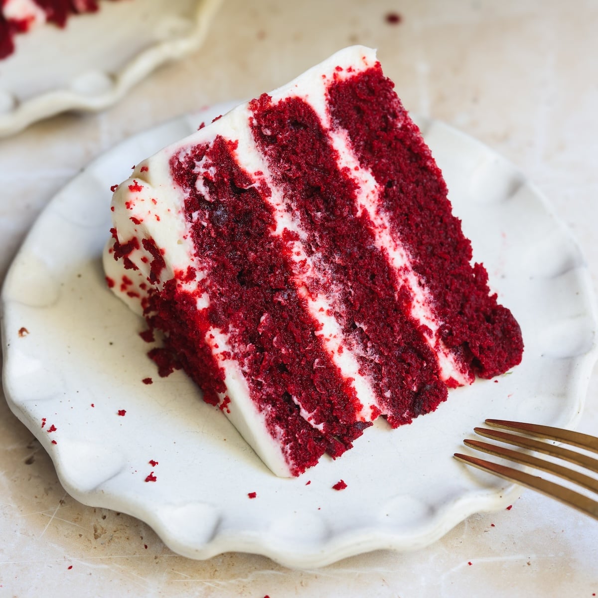 A slice of very moist and soft red velvet cake with white cream cheese frosting, served on a white plate with a gold fork beside it.