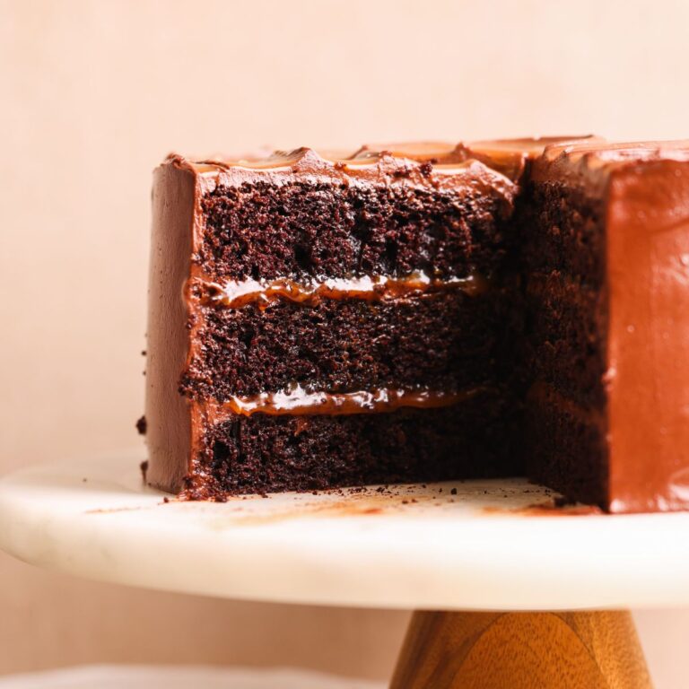 salted caramel chocolate fudge cake on a marble cake stand. cake is covered in chocolate caramel frosting and has a swirl of caramel on top. a few slices have been removed to show the soft and moist texture inside