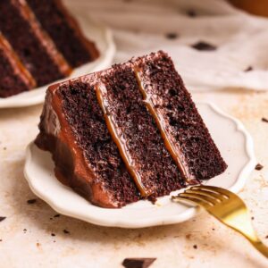 slice of salted caramel chocolate fudge cake on a small plate. cake has 3 moist layers with chocolate caramel frosting and salted caramel sauce in between.