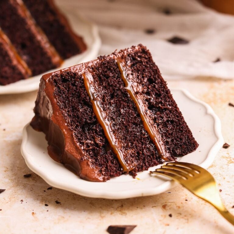slice of salted caramel chocolate fudge cake on a small plate. cake has 3 moist layers with chocolate caramel frosting and salted caramel sauce in between.