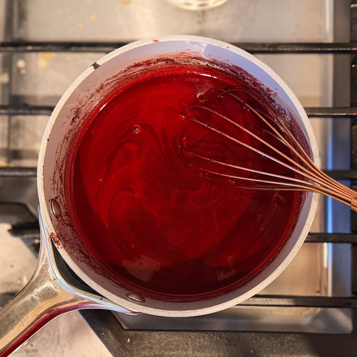 A metal saucepan with deep red mixture is being whisked on a stove, seen from above. The sauce appears smooth, and the whisk is partially submerged, swirling the mixture.