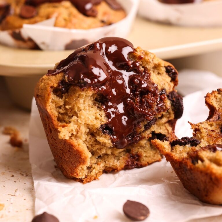 a jumbo banana chocolate muffin broken into half with more in the background. the muffin is filled with a shiny dark chocolate ganache and chocolate chips, and looks very moist in texture