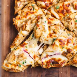 Close up of sliced chicken alfredo flatbread on a wooden cutting board, with a slice being pulled apart with a cheese pull