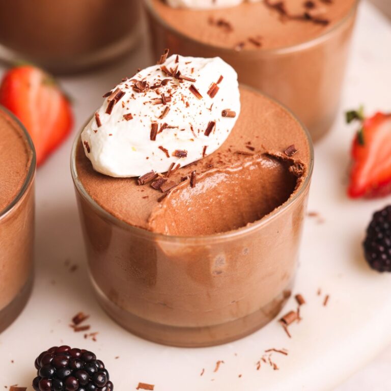 close up image of a chocolate mousse cup. it is a glass cup filled with chocolate mousse and topped with a dollop of whipped cream and grated chocolate. More cups in the background along with berries. A bite has been taken out of the mousse showing its very light, silky texture
