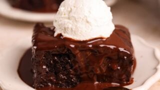 slice of chocolate sticky toffee pudding on a small plate. it is a rich and moist chocolate cake drenched in a chocolate toffee sauce with a scoop of whipped cream on top. close up picture from the side