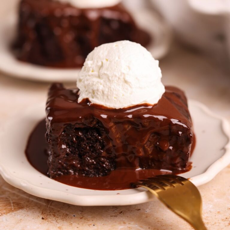 slice of chocolate sticky toffee pudding on a small plate. it is a rich and moist chocolate cake drenched in a chocolate toffee sauce with a scoop of whipped cream on top. close up picture from the side