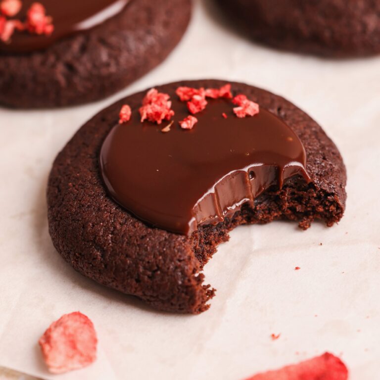 close up picture of chocolate thumbprint cookie with chocolate ganache decorated with crushed freeze dried strawberry. a bite has been taken out of the cookie