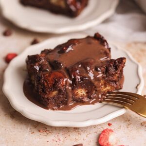 slice of chocolate bread pudding on a small plate with chocolate creme anglaise poured on top, fork resting on the side