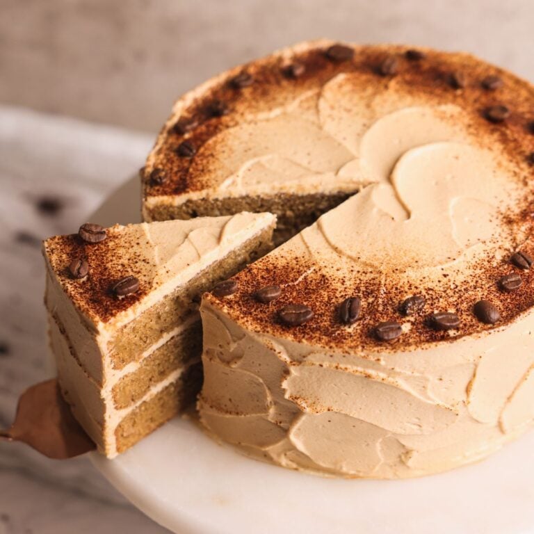 espresso cake on a white marble cake stand. cake is frosted with a coffee flavored cream cheese frosting and decorated with instant coffee powder and coffee beans. A spatula is pulling out one slice showing the 3 layers of moist cake and frosting inside