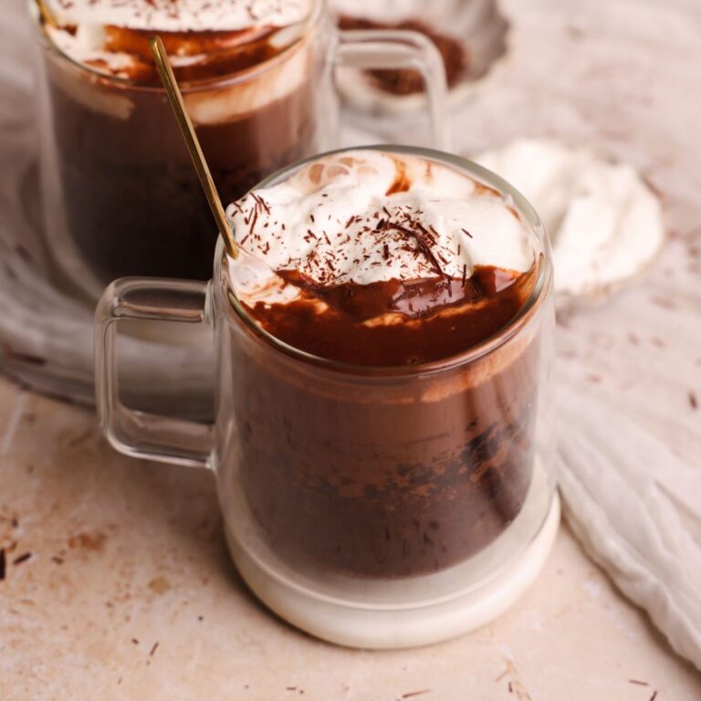 2 glass mugs with hot chocolate cakes inside. the mugs have chocolate cakes covered with hot chocolate and topped off with whipped cream and shaved chocolate. One mug is in focus