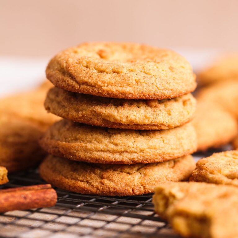 stack of 4 snickerdoodle cookies on a wire rack with more in the back. the cookies are perfectly round and thick, with a golden crispy outside coated in cinnamon sugar
