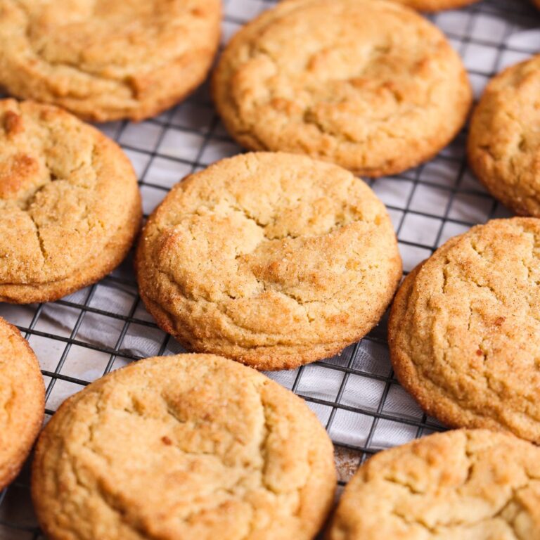 A batch of baked snickerdoodle cookies placed on a wire rack with some cinnamon sticks. The cookies look beautiful, golden and crispy and are coated with cinnamon sugar