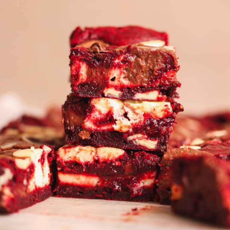 3 red velvet chocolate cheesecake brownies stacked on top of each other. they are fudgy and creamy with red brownie, chocolate cheesecake and vanilla cheesecake