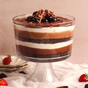 chocolate lasagna trifle in a glass trifle bowl with layers of crushed cookies, chocolate pudding and whipped cream, decorated with ganache and cherries. picture from the side