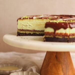 dubai chocolate cheesecake on a marble stand pictured from the side. The cheesecake has layers of chocolate crust, pistachio knafeh, chocolate ganache, and pistachio cheesecake. A few slices removed to show the creamy interior
