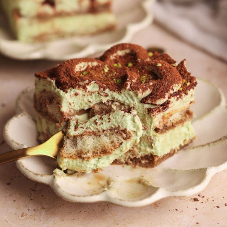 slice of pistachio tiramisu in a small plate with layers of ladyfingers and a pistachio mascarpone cream. a fork is pulling out a slice