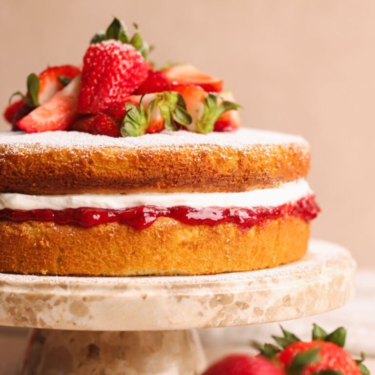 half of a victoria sandwich cake on a marble cake stand decorated with strawberries and layered with jam and whipped cream, pictured from the side