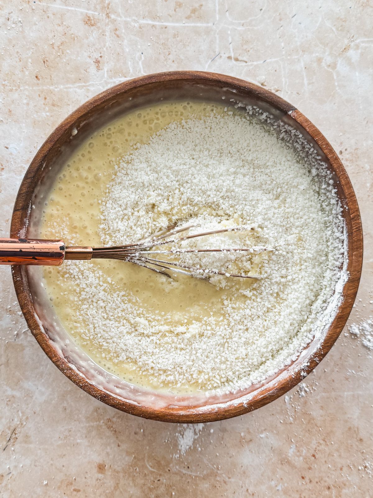 whisk mixing desiccated coconut into cake batter in a wooden bowl