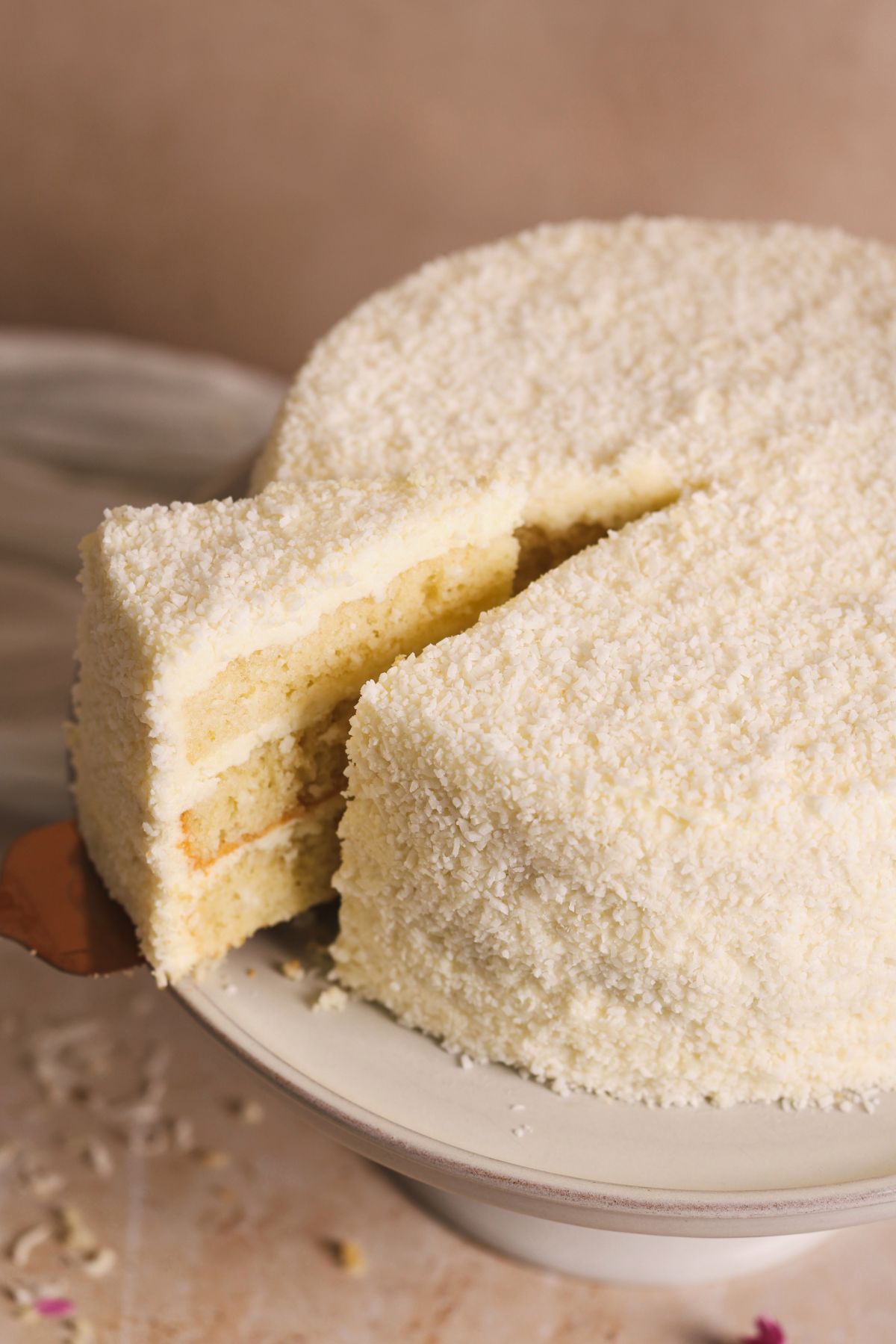 A spatula pulling a slice out of coconut cake. cake is covered with coconut cream cheese frosting and desiccated coconut