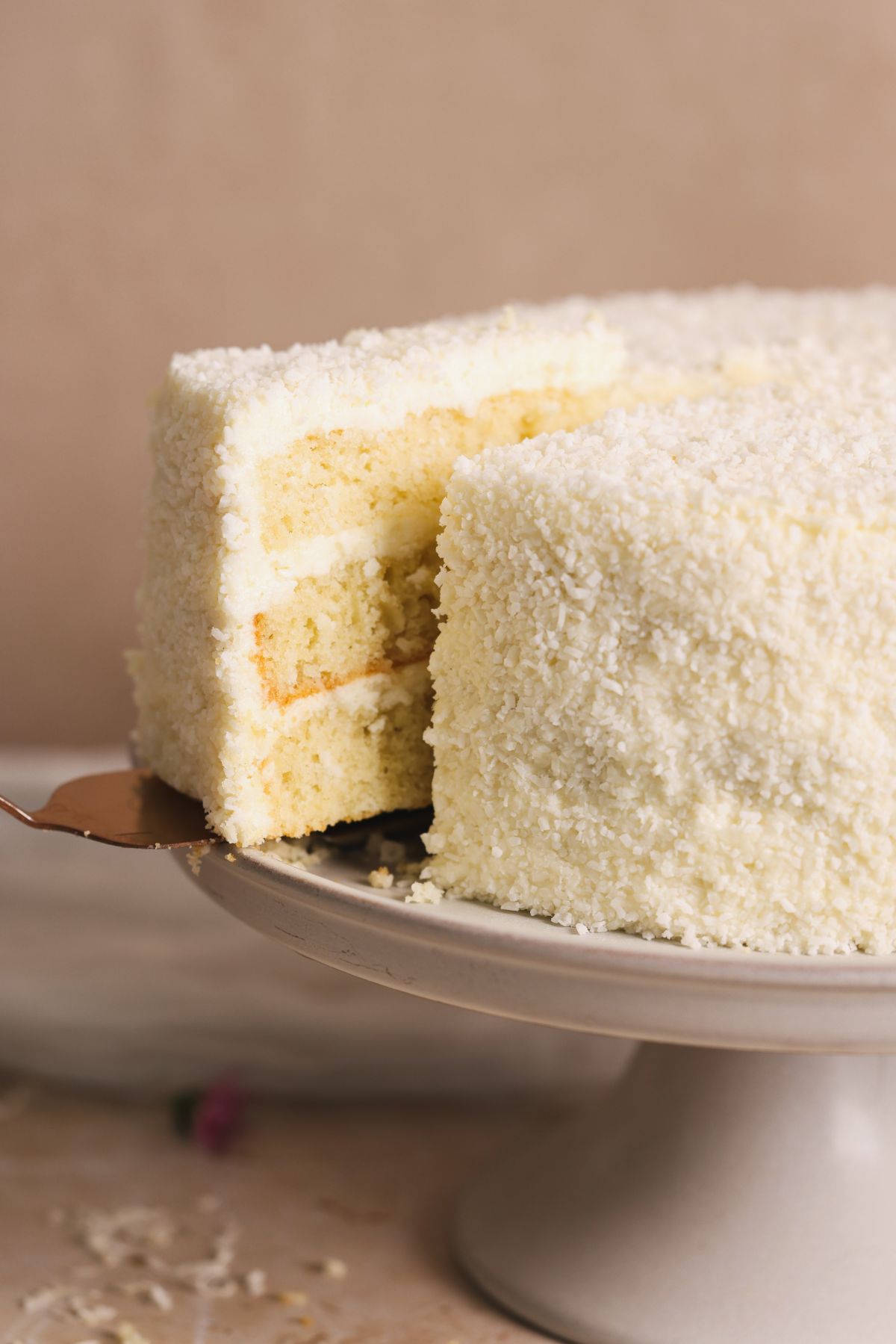 A spatula pulling a slice out of coconut cake. cake is covered with coconut cream cheese frosting and desiccated coconut, pictured from the side