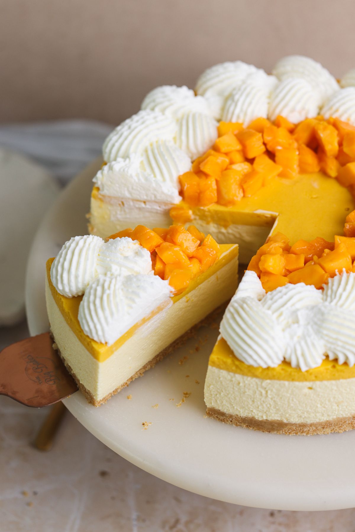 no bake mango cheesecake on a cake stand with a spatula removing a slice. the cheesecake is decorated with whipped cream and chopped mangoes