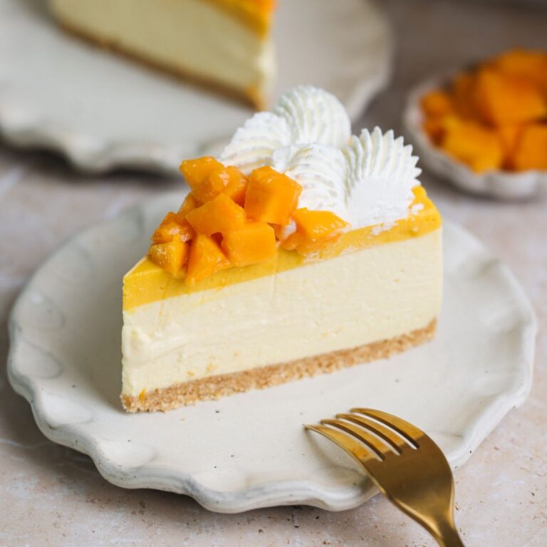 slice of no bake mango cheesecake on a small plate with another in the background. the cheesecake has a golden crust, creamy cheesecake, and mango pulp, and is decorated with mangoes and cream