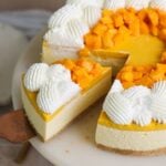 no bake mango cheesecake on a cake stand with a spatula removing a slice. the cheesecake is decorated with whipped cream and chopped mangoes