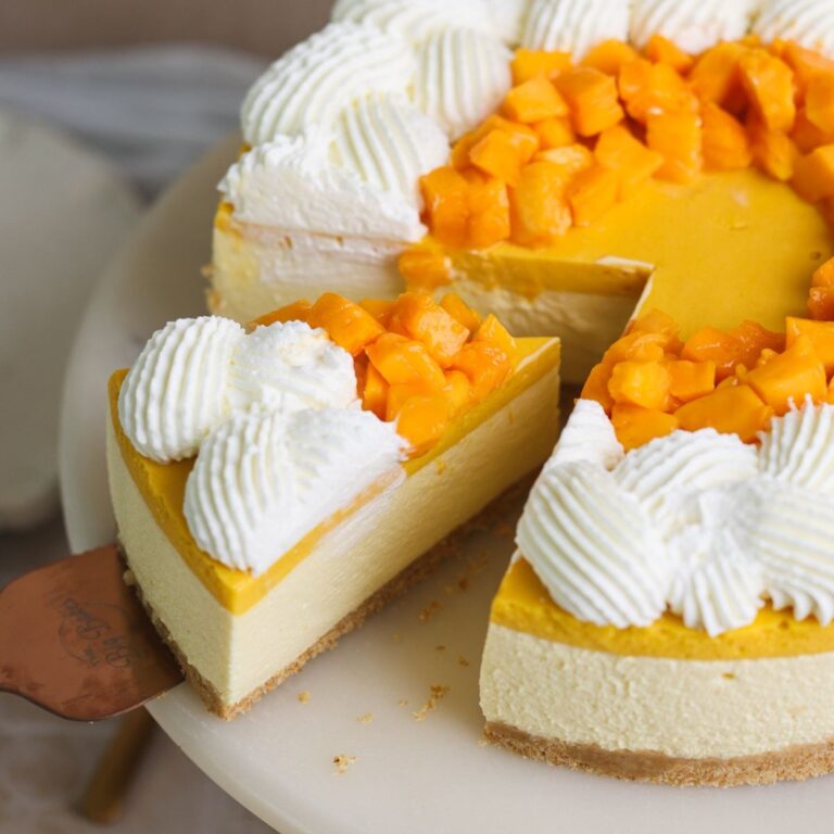 no bake mango cheesecake on a cake stand with a spatula removing a slice. the cheesecake is decorated with whipped cream and chopped mangoes
