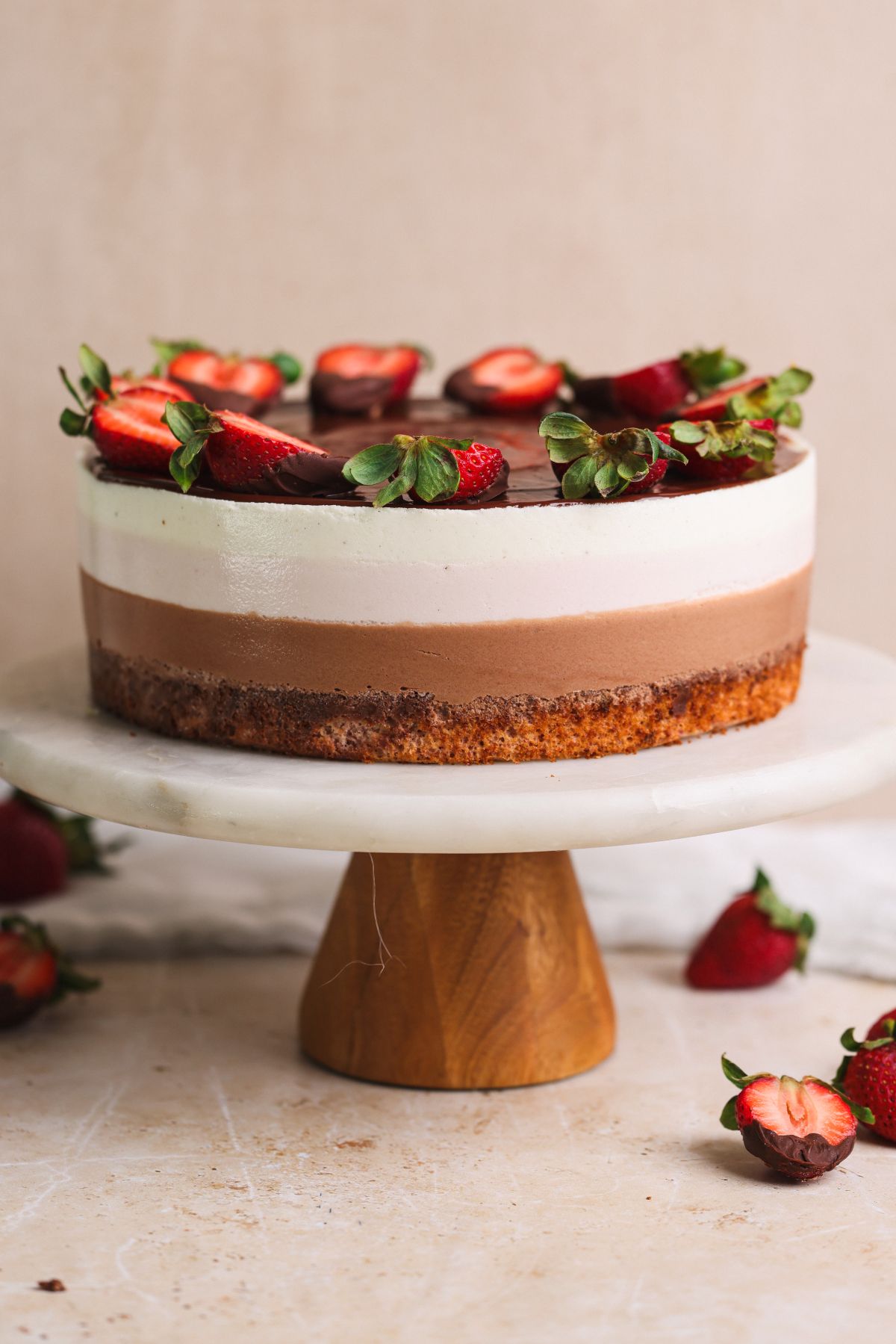 neopolitan ice cream mousse cake on a cake stand. cake has layers of chocolate, vanilla and strawberry mousse and is decorated with ganache and strawberries