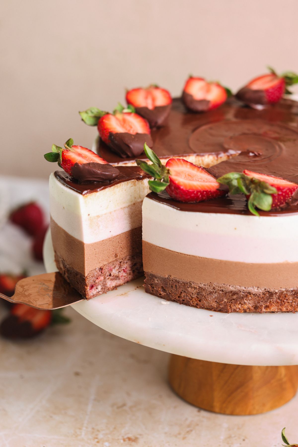 neopolitan ice cream mousse cake on a cake stand with a spatula pulling out a slice. the cake has layers of strawberry cake, chocolate mousse, strawberry mousse and vanilla mousse, and is decorated with ganache and strawberries