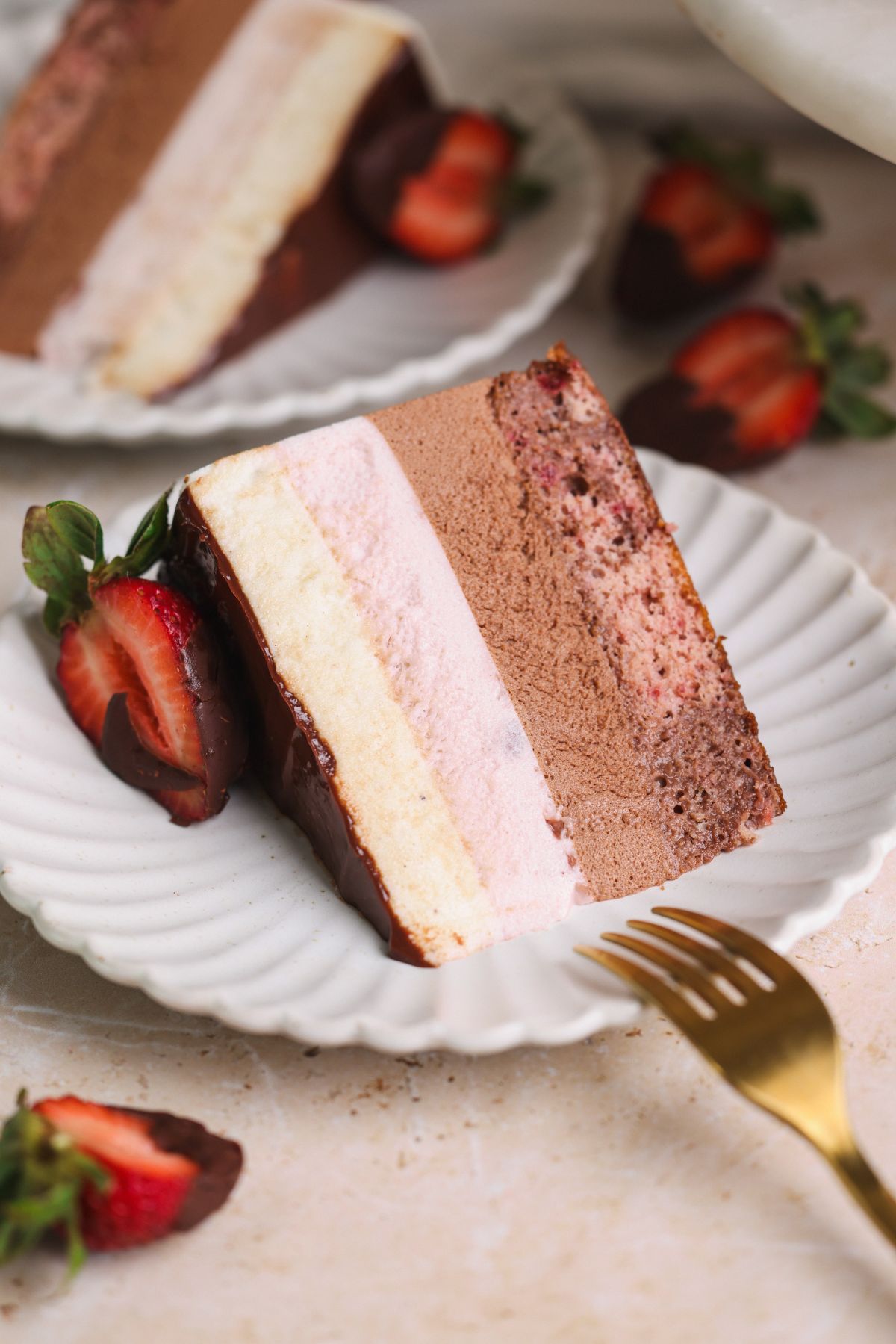 a slice of neopolitan ice cream mousse cake on a small plate with a fork resting next to it. cake has layers of chocolate mousse, strawberry mousse and vanilla mousse