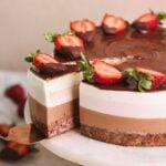 neopolitan ice cream mousse cake on a cake stand with a spatula pulling out a slice. the cake has layers of strawberry cake, chocolate mousse, strawberry mousse and vanilla mousse, and is decorated with ganache and strawberries