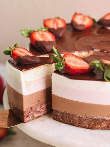 neopolitan ice cream mousse cake on a cake stand with a spatula pulling out a slice. the cake has layers of strawberry cake, chocolate mousse, strawberry mousse and vanilla mousse, and is decorated with ganache and strawberries